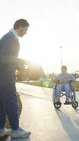 Older brother passes basketball to his younger brother, who is unable to play due to an injury and uses wheelchair. Two brothers play basketball together in park, enjoying game and spending time together. video