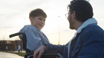 A young man in a wheelchair, who has been injured, is enjoying a conversation with his father as they relax in the park on a warm summer day. video