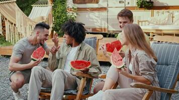 Four diverse friends laugh and share watermelon slices while relaxing in camping chairs at a scenic outdoor location during a sunny summer weekend. video