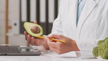 A dietitian at a clinic examines a ripe avocado while holding a pencil and reviewing materials on a laptop. Fresh vegetables are nearby, emphasizing a focus on nutrition and diet. video
