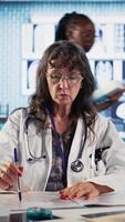 Vertical Senior doctor and patient discussing diagnostic results in a modern medical office, showcasing advanced technology using records files displaying x rays and MRI scans. Care management. Camera A. video