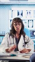 Vertical Senior woman evaluating diagnostic results with her medic in a cabinet, using advanced tech tools to give the correct results to his patient. Care management within medical industry. Camera A. video
