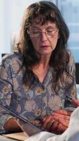 Vertical Senior woman evaluating diagnostic results with her medic in a cabinet, using advanced tech tools to give the correct results to his patient. Care management within medical industry. Camera B. video