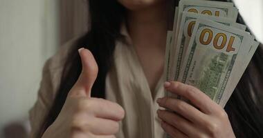 woman holds multiple dollar bills in her hands while smiling and giving thumbs up She is indoors surrounded warm lighting creates pleasant atmosphere video