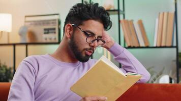 indiano homem lendo interessante livro girando Páginas sorridente desfrutando literatura levando uma descansar em sofá video