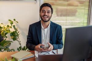 Smiling indian businessman in suit use phone while working from home office and looking at camera photo