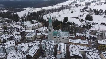 Aerial pan of Cortina d'Ampezzo in winter, featuring the Basilica Minore's green spire, snow covered fields, forests, and a visible construction crane. video