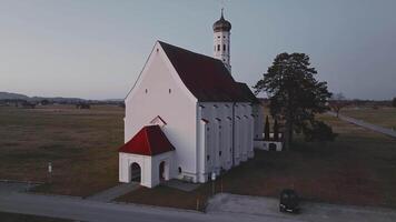 Aerial view of a white church with a red roof and onion shaped dome, surrounded by open fields, snow capped mountains, and warm sunset lighting. video