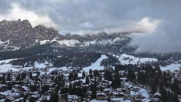 Aerial view of Cortina d'Ampezzo with snow covered rooftops, winding roads, evergreen trees, and Dolomite peaks partially shrouded in mist. video