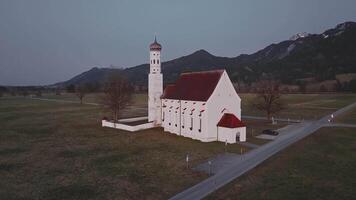 Aerial view of a white church with a red roof and tall tower, surrounded by fields, roads, and distant snow capped mountains in soft evening light. video