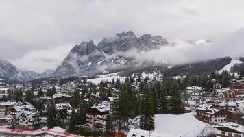 Aerial view of Cortina d'Ampezzo with snow covered rooftops, the Basilica Minore bell tower, and dramatic Dolomite peaks under low clouds. video