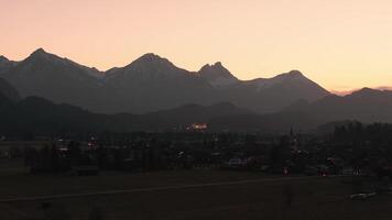 Antenne Aussicht von neuschwanstein Schloss beleuchtet beim Sonnenuntergang, mit das bayerisch Alpen und ein Kirche im das rahmen. das Himmel Übergänge zu Dämmerung. video