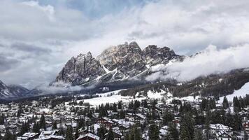 Aerial view of Cortina d'Ampezzo with snow covered rooftops, dense forests, and Dolomites peaks partially shrouded in drifting clouds. video