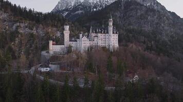 Aerial view of Neuschwanstein Castle at sunset, surrounded by dense forest and snow capped mountains, with a small church in the foreground. video