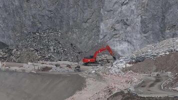 An aerial view of a mining site in the Alps shows an orange excavator breaking rocks, creating debris piles, and operating amidst steep rocky cliffs. video