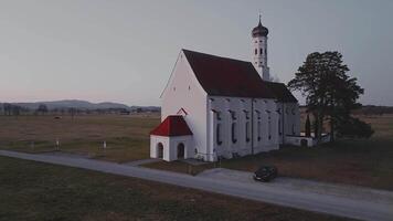 Antenne Aussicht von ein Weiß Kirche mit ein rot Dach und hoch Turm, umgeben durch öffnen Felder und ein gepflastert Straße, mit glatt Kamera Bewegung beim Sonnenuntergang. video