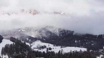 Aerial view of snow covered Dolomite mountains, dense forests, and ski trails. Low clouds drift, revealing rugged peaks and adding dynamic motion. video