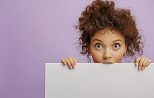 Young Woman With Curly Hair Peeks Over a Blank Canvas Against a Purple Background in a Playful Expression photo