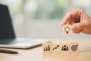 Hand Placing Wooden Block With Light Bulb Icon Above Other Blocks on Desk With Laptop in Background photo