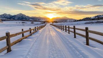 A snowy road leads to a wooden fence with the sun setting behind it photo