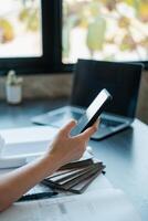 Close-up of a hand holding a smartphone at a desk with a laptop and documents, in a well-lit office setting. photo