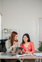 Two women discussing work in a bright office, using a tablet and surrounded by documents, showcasing teamwork and technology. photo