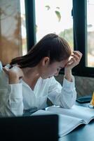 Businesswoman in white shirt appears stressed while working at desk with open book and laptop in a bright office. photo