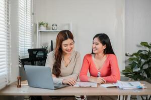 Two women working together at a home office desk, smiling and discussing documents with a laptop in a bright, modern setting. photo