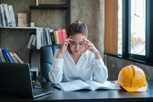 A female engineer appears stressed while working at her desk, surrounded by books and a safety helmet in a modern office environment. photo