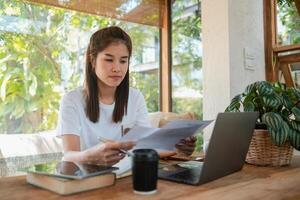 A young woman focuses on her work, surrounded by greenery and natural light, using a laptop and reviewing documents in a cozy home setting. photo