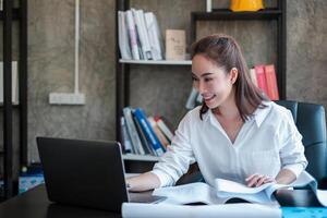 A woman in a modern office setting, working on a laptop, surrounded by books and documents, smiling and focused. photo
