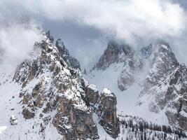 Snow covered peaks of the Dolomites in Italy, featuring jagged rock formations, mist, clouds, and sparse trees near the rugged terrain's base. photo