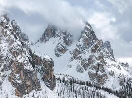 Snow covered Dolomite peaks near Cortina, Italy, partially shrouded in clouds. Sparse coniferous trees dot the lower slopes, contrasting the rugged terrain. photo