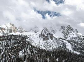 Snow covered peaks of the Dolomite Mountains in Italy rise above dense evergreen forests, with clouds partially shrouding the rugged terrain. photo