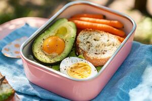 A pink lunch box with an avocado, carrot, and egg photo
