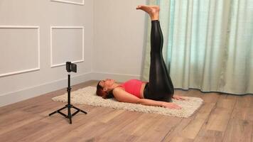 middle aged woman doing yoga lying on the floor at home and also broadcasting to her followers using a smartphone mounted on a tripod video