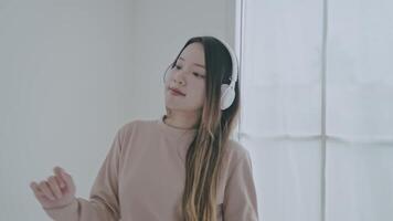 A young woman wearing white headphones smiles while listening to music on a comfortable sofa. She stretches her arms, enjoying a peaceful moment in a bright, inviting living space. video