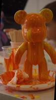 A bright orange figurine in the shape of a bear, covered with fresh paint, dries while the artist is engaged in another statuette, but with similar patterns, but in a different color scheme. video