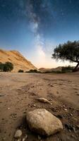 A serene desert scene features rolling sand dunes and a prominent rocky foreground. The twilight sky reveals distant stars, enhancing the tranquil atmosphere while sparse vegetation adds depth photo