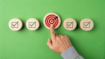 Hand pointing at red bullseye target among wooden checkmark blocks on green background, representing focus, goal setting, and achievement photo