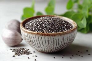 Chia seeds in a decorative bowl with fresh herbs on a kitchen countertop. Ceramic bowl filled with black chia seeds on clean light surface for healthy desserts in culinary setting photo