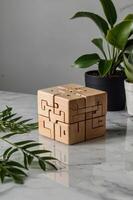 Wooden puzzle cube displayed on a marble surface surrounded by green plants in a bright indoor setting photo