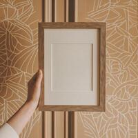 A person holds up an empty, wooden picture frame against a patterned brown and white background photo