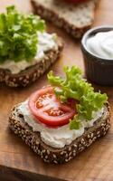 A slice of multigrain bread topped with cream cheese, tomato, and lettuce sits on a wooden surface, accompanied by a small bowl of cream cheese photo