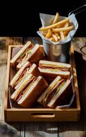A wooden tray holds several neatly arranged sandwiches featuring layers of meat and cheese, accompanied by a metal container of golden French fries photo