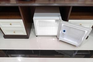 mini fridge in low angle view, inside it s empty shelves placed under wooden counter and next to wood closet and chair in a luxury hotel room.1 photo
