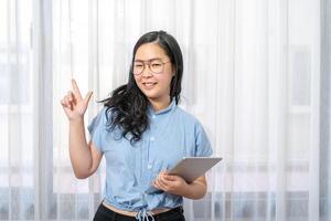 Happy Asian woman fingering up and pointing with tablet computer on her hand at copy space while standing in front of the transparent curtain see through the window light.aa photo
