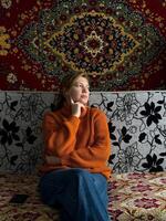 In a traditional Russian home, a woman wearing a cozy orange sweater sits on a patterned couch, with a richly decorated vintage carpet displayed on the wall behind her. photo