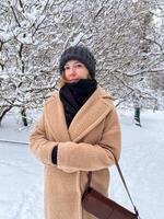 In a snowy park, a woman in a cozy beige coat, black scarf, and knitted hat stands gazing into the distance. Snow-covered branches of nearby trees create a serene winter atmosphere. photo