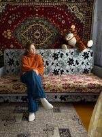 A woman in a cozy orange sweater sits on a patterned couch in a traditional Russian home, with a richly decorated vintage carpet hanging behind her. photo